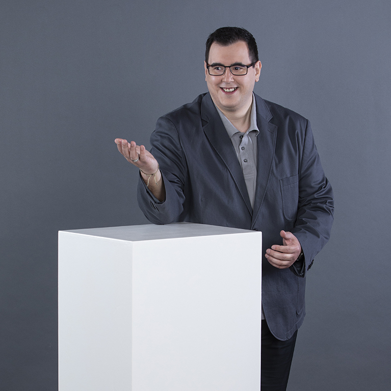 ein lächelnder Mann mit Brille in einem dunklen Sakko über einem hellgrauen Poloshirt lehnt sich mit einem Arm auf einen weißen Quader und gestikuliert mit der anderen Hand vor einem grauen Hintergrund 