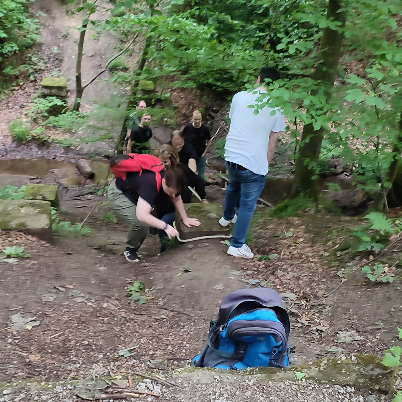 Gruppe von Wanderern, die einen felsigen Weg in einem Wald hinaufklettern, wobei im Vordergrund ein blauer Rucksack auf dem Boden liegt