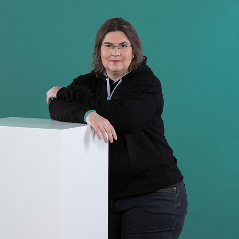 lächelnde Frau mit Brille steht vor einem petrolfarbigen Hintergrund, trägt einen schwarzen ICN-Hoodie und eine dunkle Hose und lehnt sich mit verschränkten Armen auf einen weißen Block 