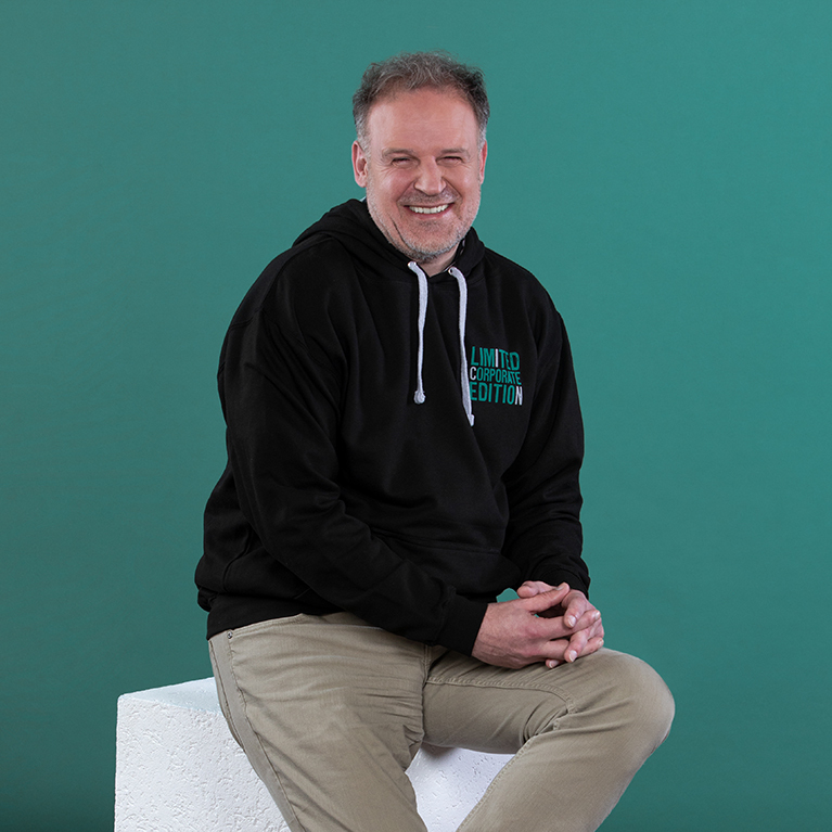 lächelnder Mann mit Bart und lockeren Haaren sitzt in einem schwarzen ICN-Hoodie und einer beigen Hose auf einem weißen Block vor einem petrolfarbigen Hintergrund 