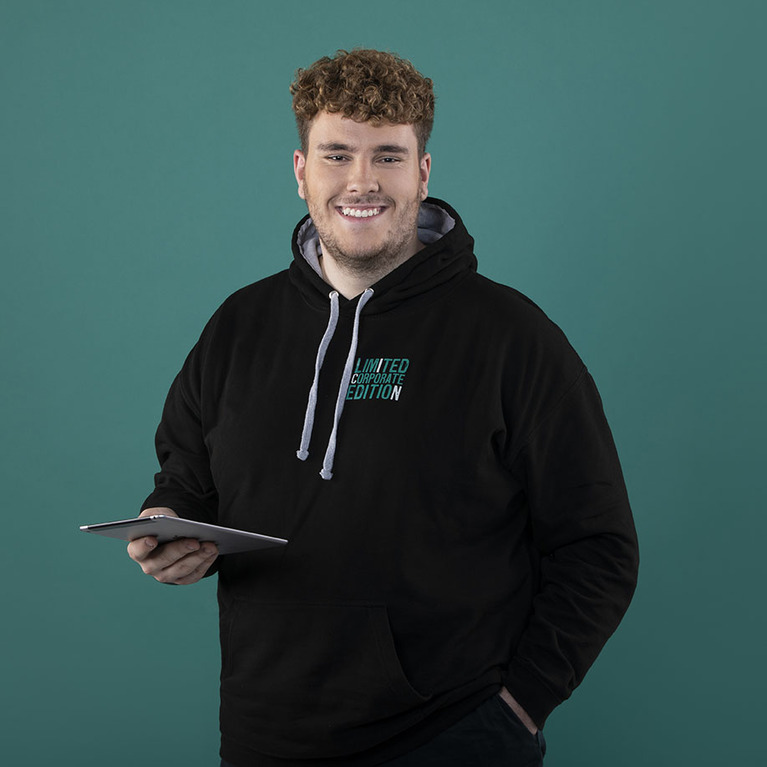 lächelnder Mann mit lockigen Haaren in einem schwarzen ICN-Hoodie hält ein Tablet in der Hand und hat die andere Hand in der Hosentasche vor petrolfarbigen Hintergrund  