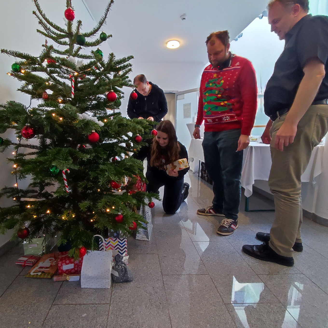 vier Personen, die in einem Flur einen Weihnachtsbaum mit Geschenken darunter schmücken 