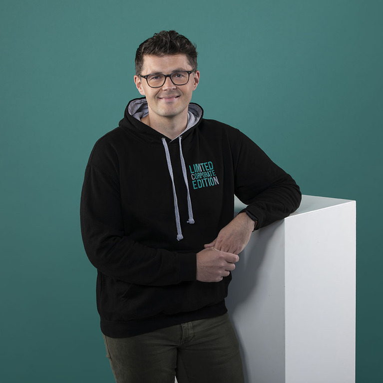 lächelnder Mann mit Brille in einem schwarzen ICN Hoodie und olivgrüner Hose lehnt sich mit verschränkten Armen auf einen weißen Würfel vor petrolfarbigen Hintergrund 