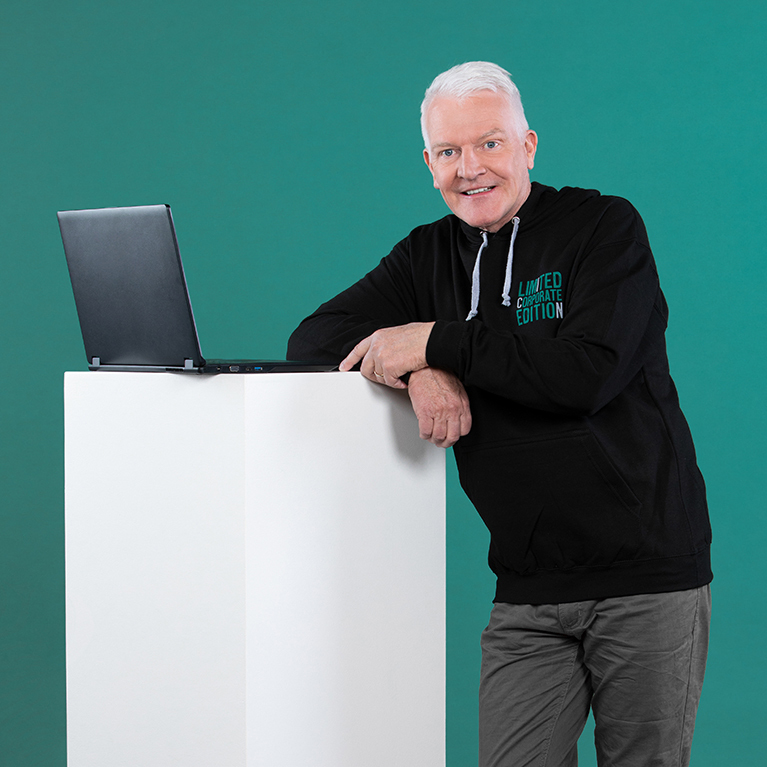 älterer lächelnder Mann steht in einem schwarzen ICN-Hoodie und dunkler Hose vor einem petrolfarbigen Hintergrund, wobei er sich mit den Armen auf einen weißen Würfel stützt, auf dem ein Laptop liegt