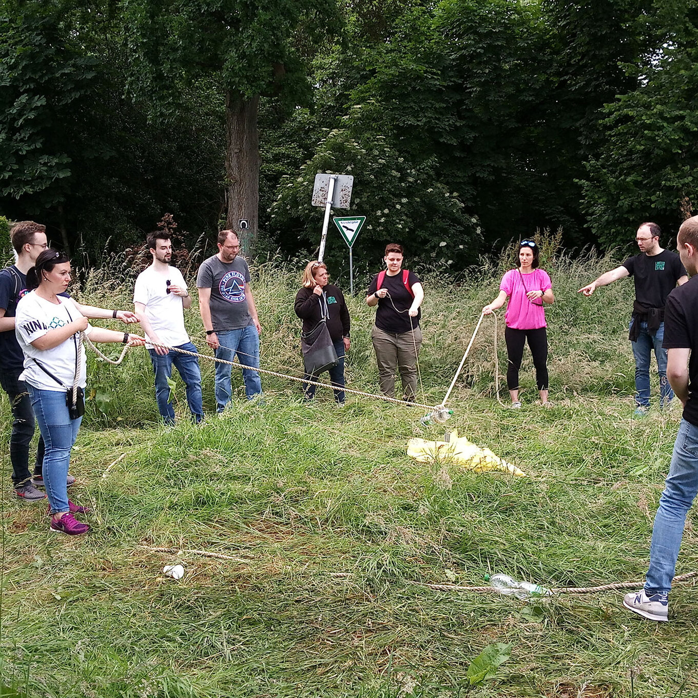 Gruppe von etwa 9 Personen, die an einem Seil ziehen, das durch eine Wiese verläuft, im Hintergrund sind Bäume zu sehen 
