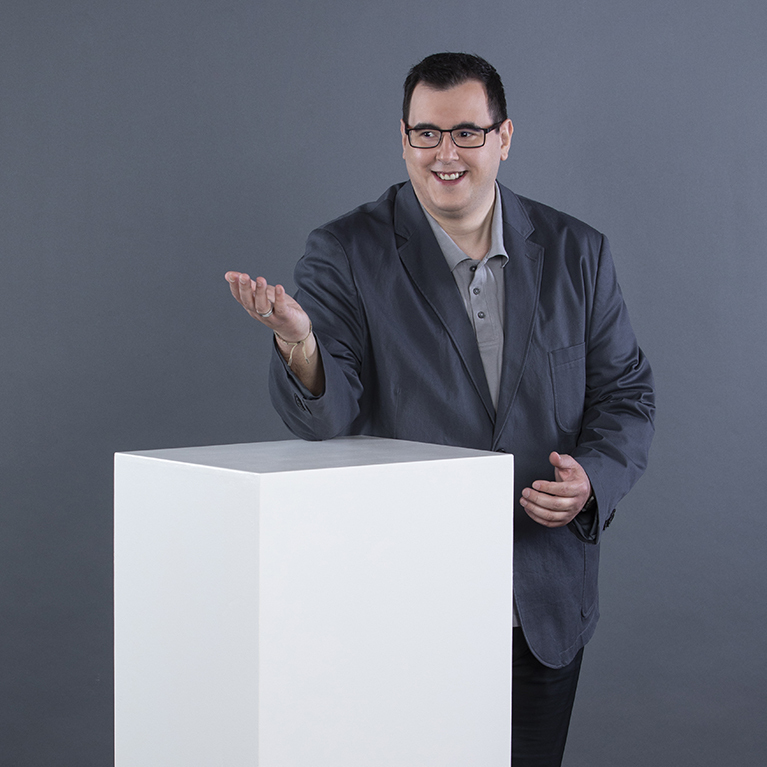 ein lächelnder Mann mit Brille in einem dunklen Sakko über einem hellgrauen Poloshirt lehnt sich mit einem Arm auf einen weißen Quader und gestikuliert mit der anderen Hand vor einem grauen Hintergrund 