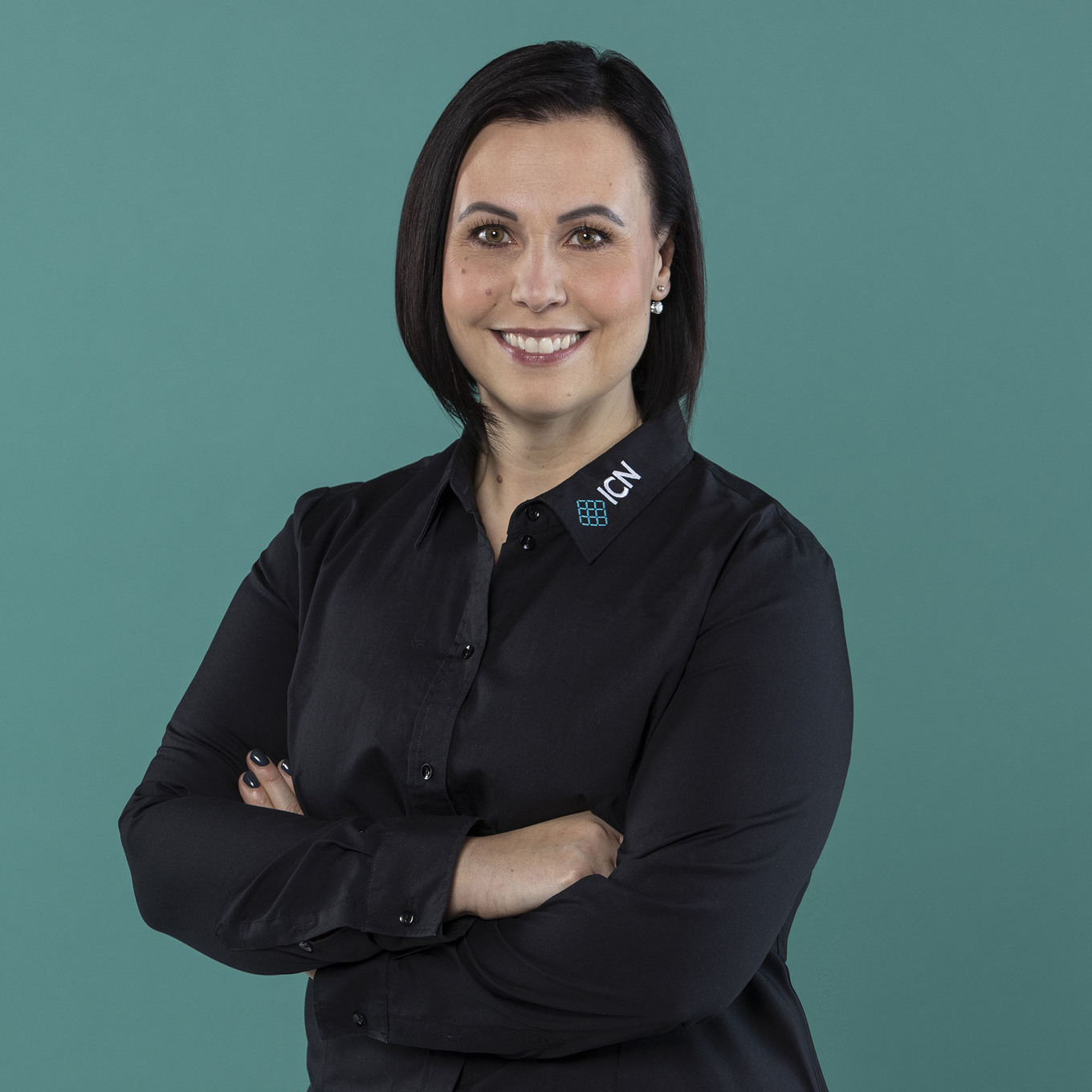 lächelnde Frau mit kurzen, dunklen Haaren steht vor einem petrolfarbigen Hintergrund, trägt ein schwarzes ICN-Hemd und hat die Arme verschränkt 