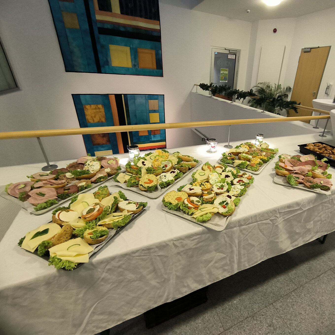 Buffet in einem Innenraum, auf einem langen Tisch, der mit einem weißen Tischtuch bedeckt ist, stehen mehrere Platten mit kleinen Sandwiches, Häppchen und Salaten