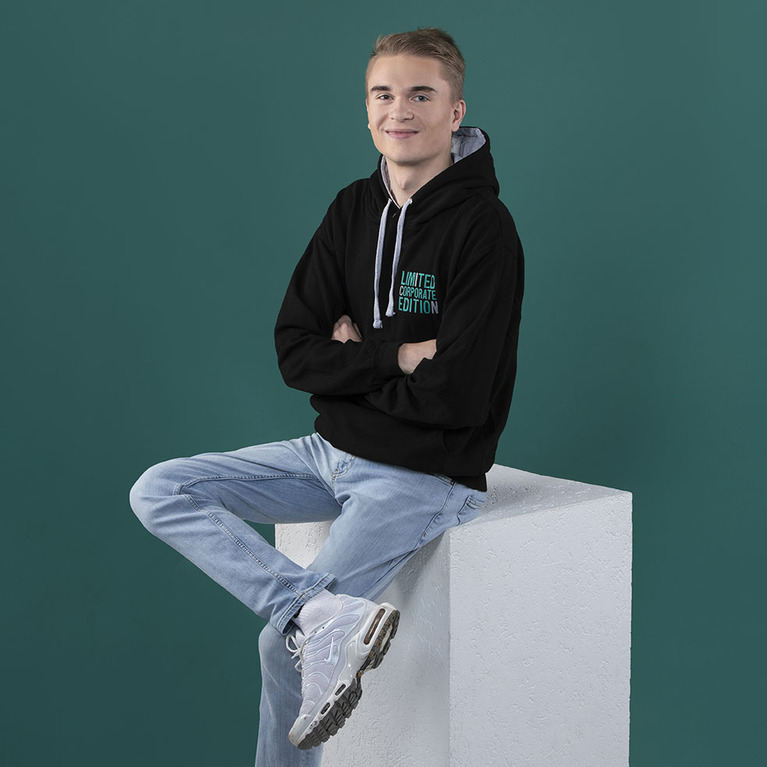 Mann in einem schwarzen ICN-Kapuzenpullover und Jeans, er sitzt auf einem weißen Würfel, verschränkt die Arme und lächelt in die Kamera, der Hintergrund ins Petrol