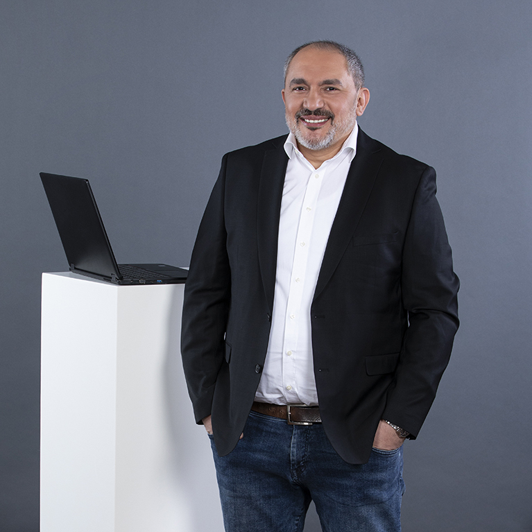 ein lächelnder Mann mit Bart steht in einem schwarzen Sakko über einem weißen Hemd und einer Jeans vor einem grauen Hintergrund, er hat die Hände in den Hosentaschen und lehnt neben einem weißen Quader, auf dem ein Laptop steht 