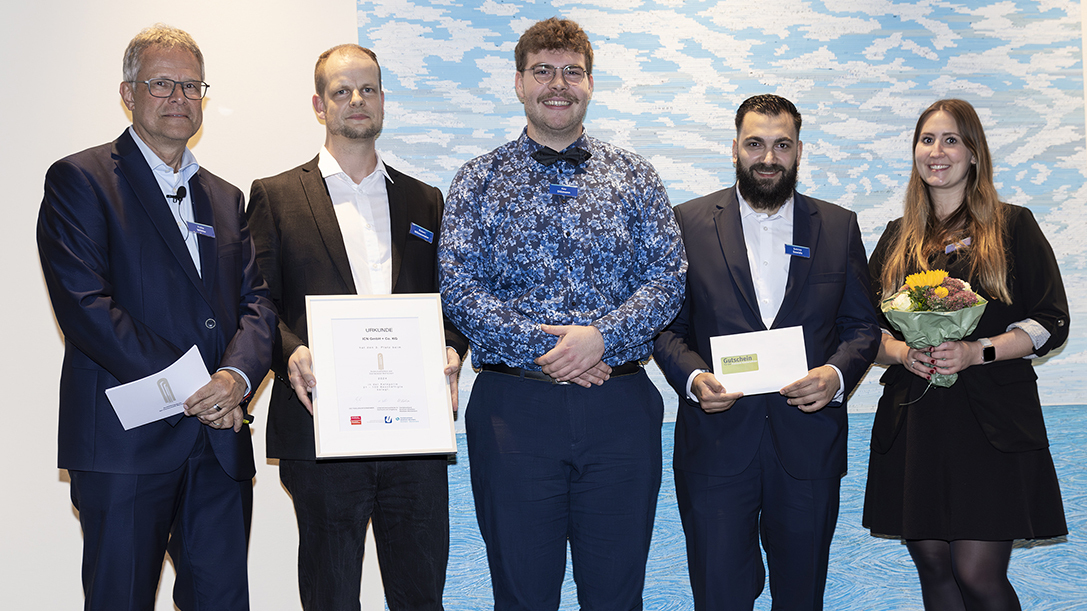 fünf Personen bei der Verleihung des Ausbildungspreises. Zwei Männer halten eine Urkunde, ein weiterer Mann hält ein Umschlag, eine Frau hält einen Blumenstrauß