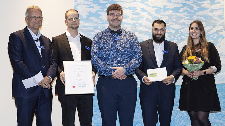 fünf Personen bei der Verleihung des Ausbildungspreises. Zwei Männer halten eine Urkunde, ein weiterer Mann hält ein Umschlag, eine Frau hält einen Blumenstrauß