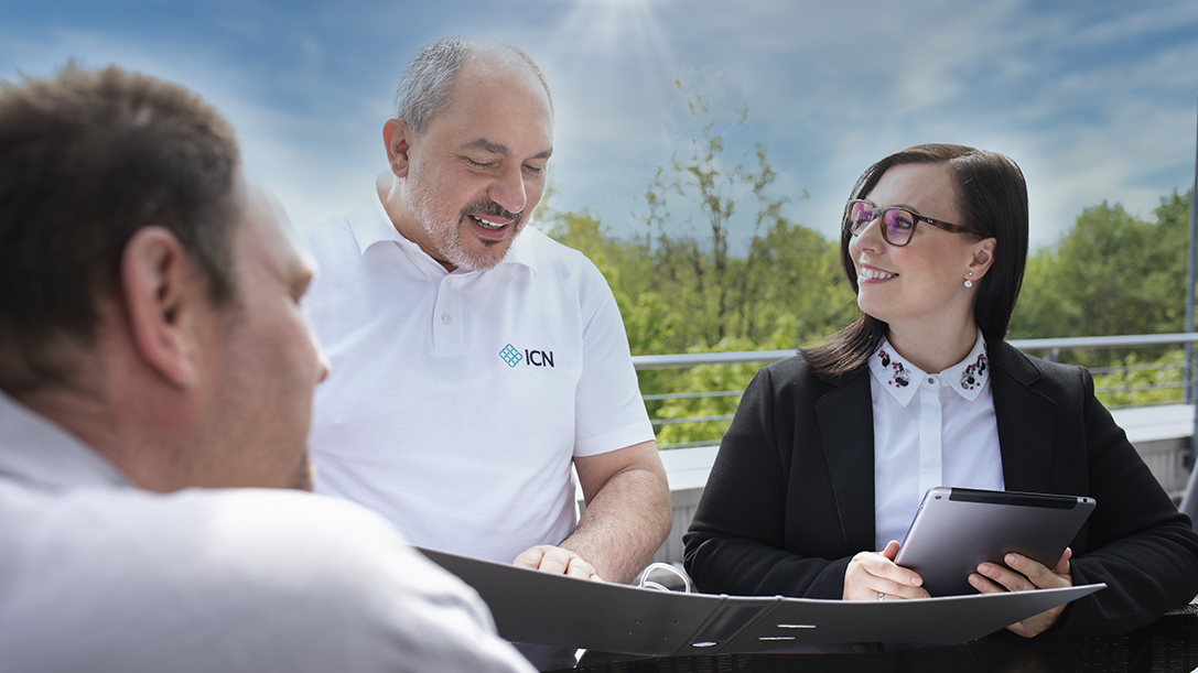 ein älterer Mann in einem weißen Poloshirt, eine Frau mit Brille in einem schwarzen Blazer mit einem Tablet und ein Mann im Vordergrund, von hinten sichtbar, sitzen an einem Tisch im Freien bei Sonnenschein und unterhalten sich 