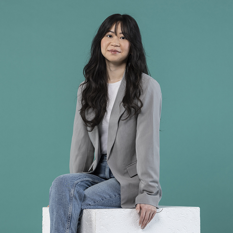 lächelnde junge Frau mit langen, lockigen Haaren sitzt in einem grauen Blazer über einem weißen Oberteil und einer Jeans auf einem weißen Block vor einem petrolfarbigen Hintergrund 