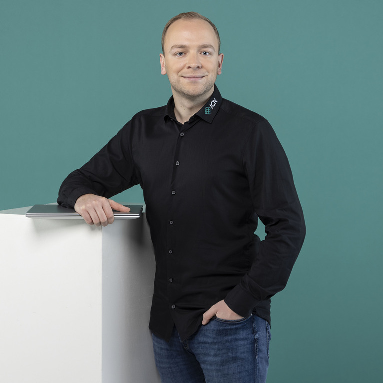 lächelnder Mann mit kurzen Haaren steht in einem schwarzen Hemd und einer Jeans vor einem petrolfarbigen Hintergrund, wobei er sich mit der rechten Hand auf einen weißen Würfel stützt