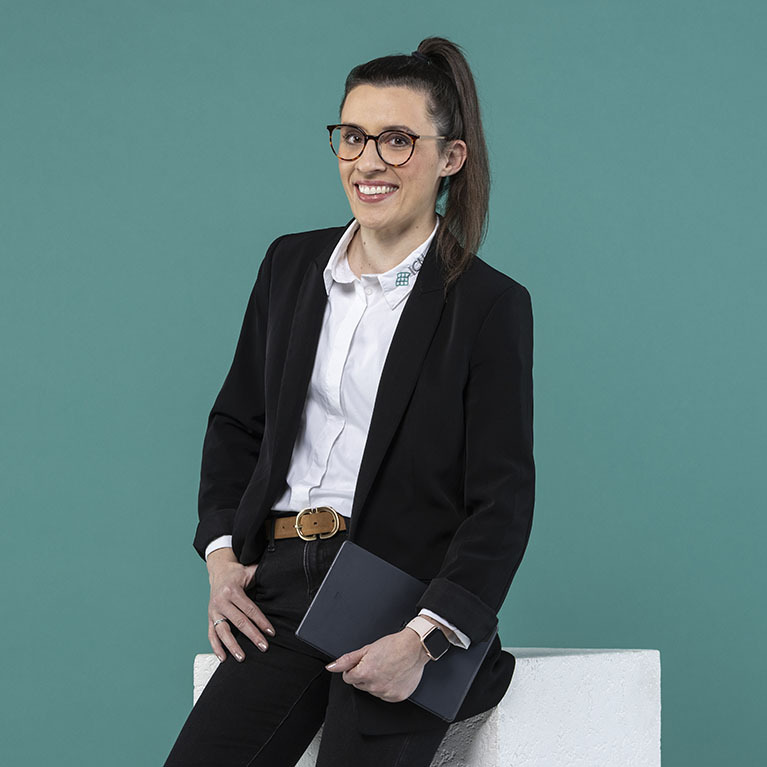 lächelnde Frau mit Brille und einem Pferdeschwanz steht in einem schwarzen Blazer über einer weißen Bluse vor einem petrolfarbigen Hintergrund, hält ein Notebook und lehnt sich leicht an einen weißen Gegenstand 