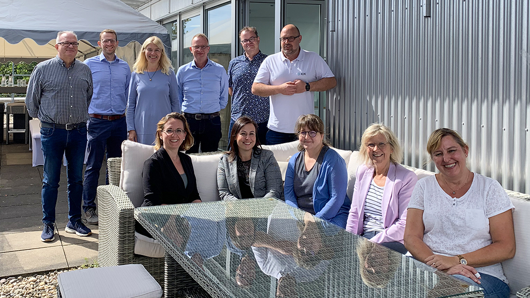 Elf Personen posieren für ein Gruppenfoto auf einer Außenterrasse vor einer Metallfassade, wobei einige auf weißen Sofas sitzen und andere dahinter stehen 