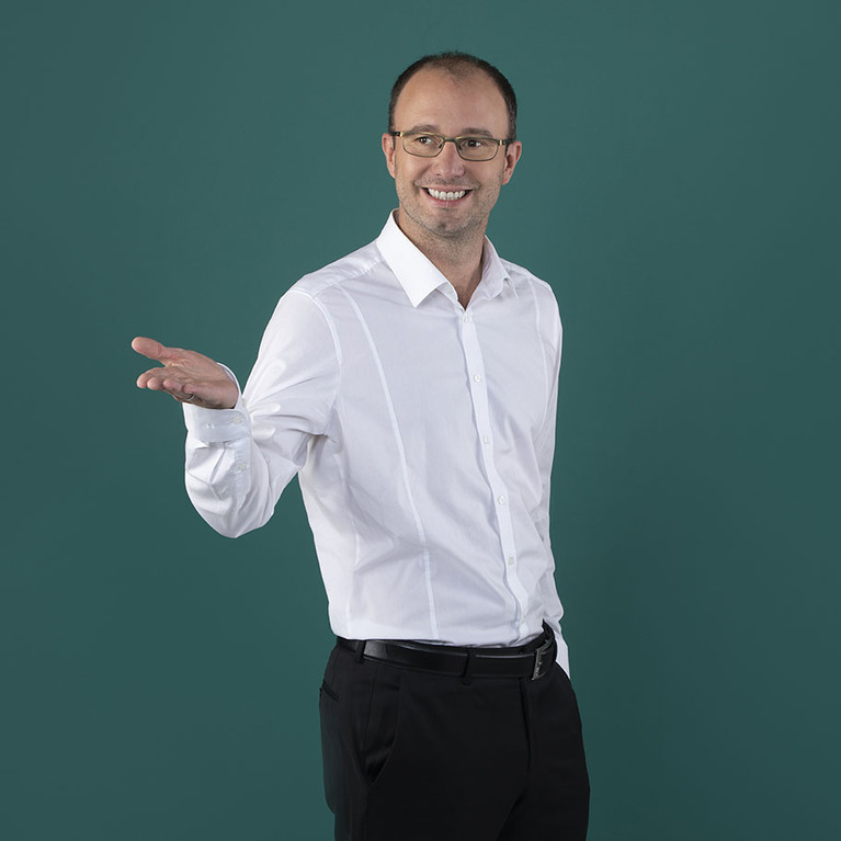 Mann in einem weißen Hemd und schwarzer Hose, trägt eine Brille, blickt lächelnd in die Kamera und streckt eine Hand aus, steht vor petrolfarbigen Hintergrund 