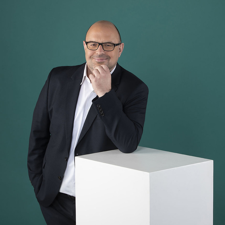 lächelnder Mann mit Brille und Anzug lehnt sich mit einem Arm auf einen weißen Würfel, hat die andere Hand am Kinn und blickt zur Seite, steht vor einem petrolfarbigen Hintergrund