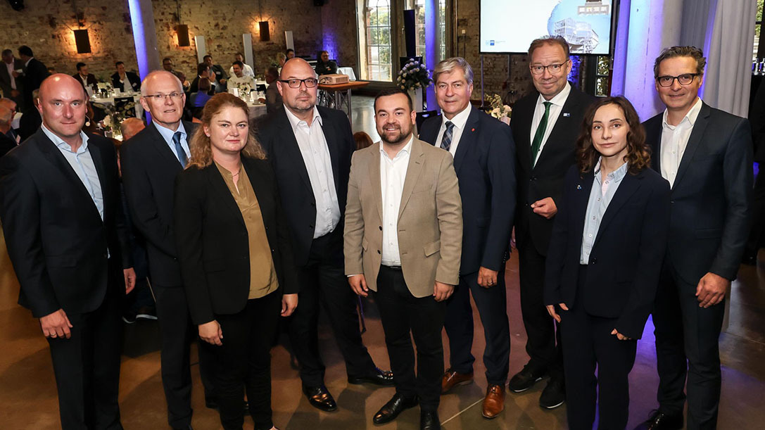 Neun Personen, überwiegend in dunkler Business-Kleidung, stehen für ein Gruppenfoto in einem festlich beleuchteten Innenraum mit blauen Lichtern 