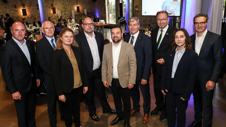 Neun Personen, überwiegend in dunkler Business-Kleidung, stehen für ein Gruppenfoto in einem festlich beleuchteten Innenraum mit blauen Lichtern 