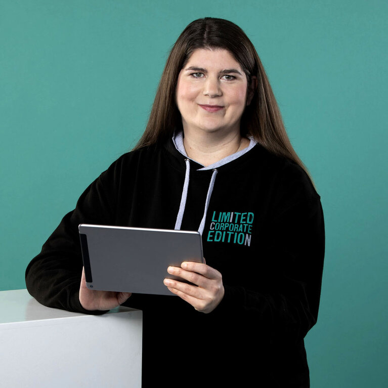lächelnde Frau mit langen, dunklen Haaren trägt einen schwarzen ICN-Hoodie, hält ein Tablet in den Händen und lehnt sich leicht an einen weißen Block vor einem petrolfarbigen Hintergrund 