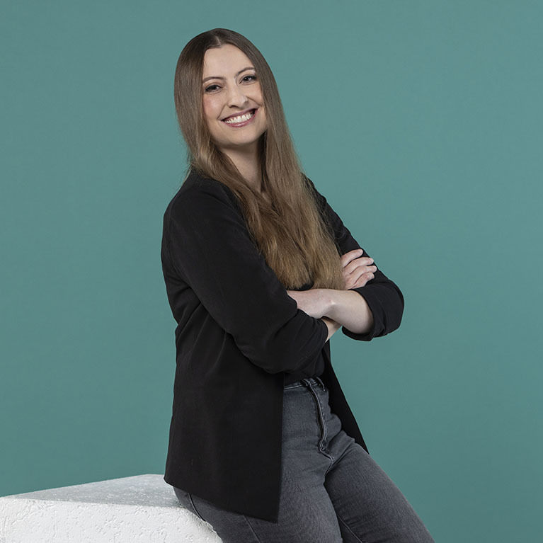 lächelnde Frau mit langen, hellbraunen Haaren sitzt auf einem weißen Block vor einem petrolfarbigen Hintergrund, trägt einen schwarzen Blazer und dunkle Jeans und hat die Arme verschränkt 
